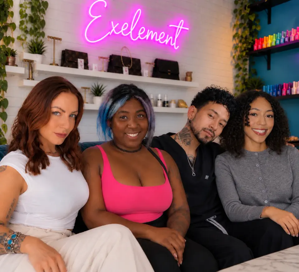 Four Exelement Beauty team members seated beneath the studio’s pink neon sign